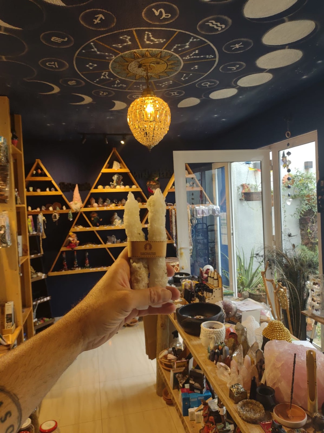Interior da loja SacraZen, com teto azul pintado de luas e signos, lustre âmbar e prateleiras de madeira com cristais